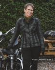 Person wearing a plaid jacket standing next to a motorcycle with a helmet on a table in the background.