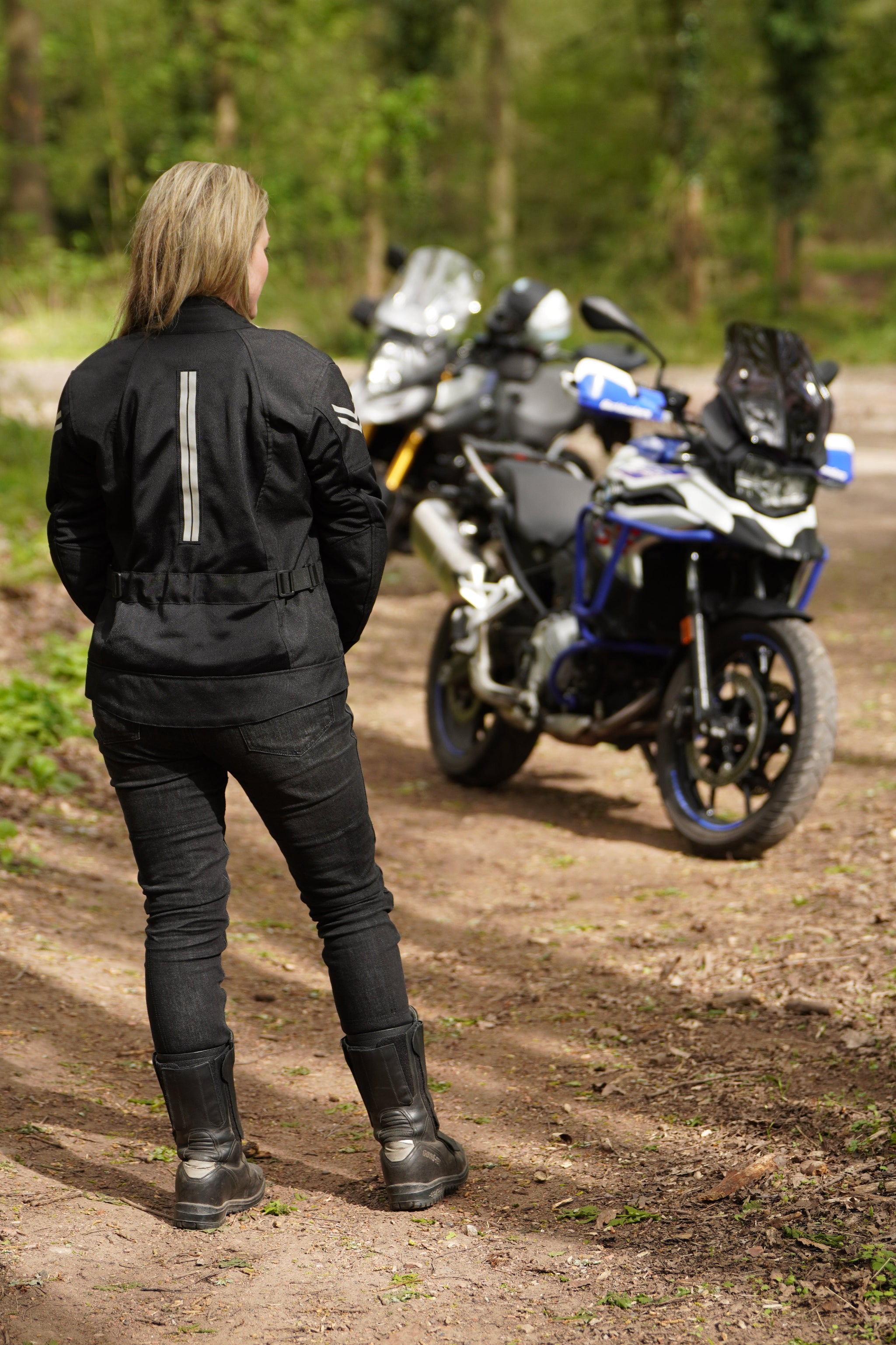 a blond woman standing back to the camera by her big motorcycle wearing a black mesh motorcycle jacket for women