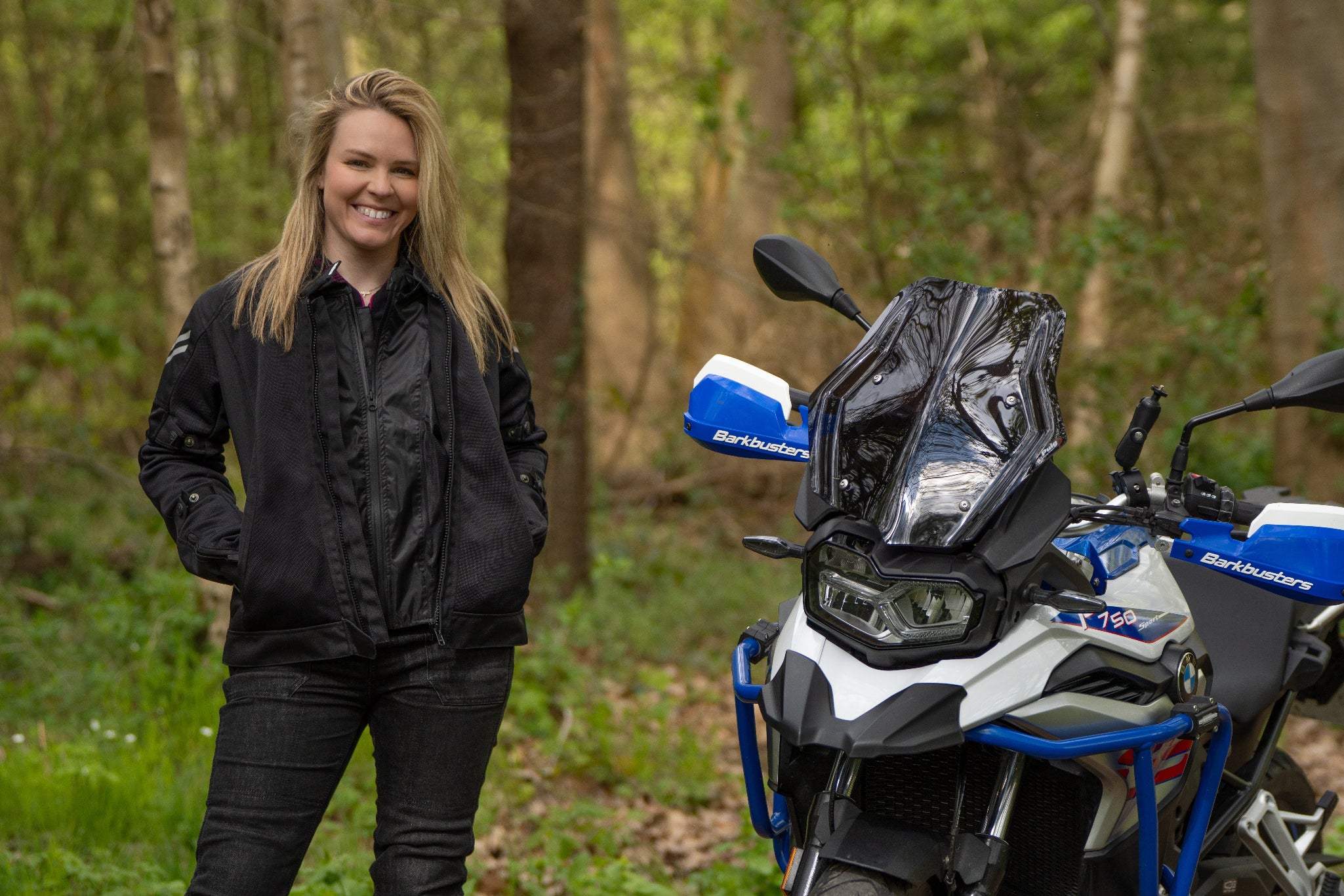 a blong smiling woman standing by her big motorcycle wearing a black mesh motorcycle jacket for women