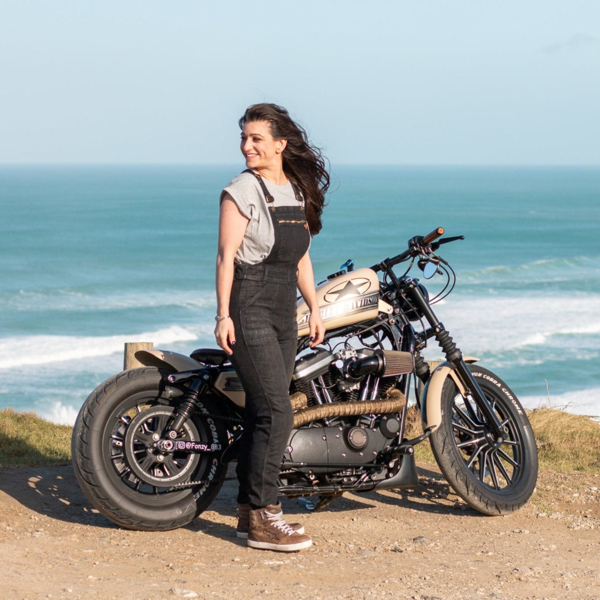 A woman by the beach with her motorcycle wearing Black women's motorcycle overall from Moto Girl