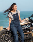 A woman by the beach with her motorcycle wearing blue women's motorcycle overall from Moto Girl with zipper pocket detail on the chest