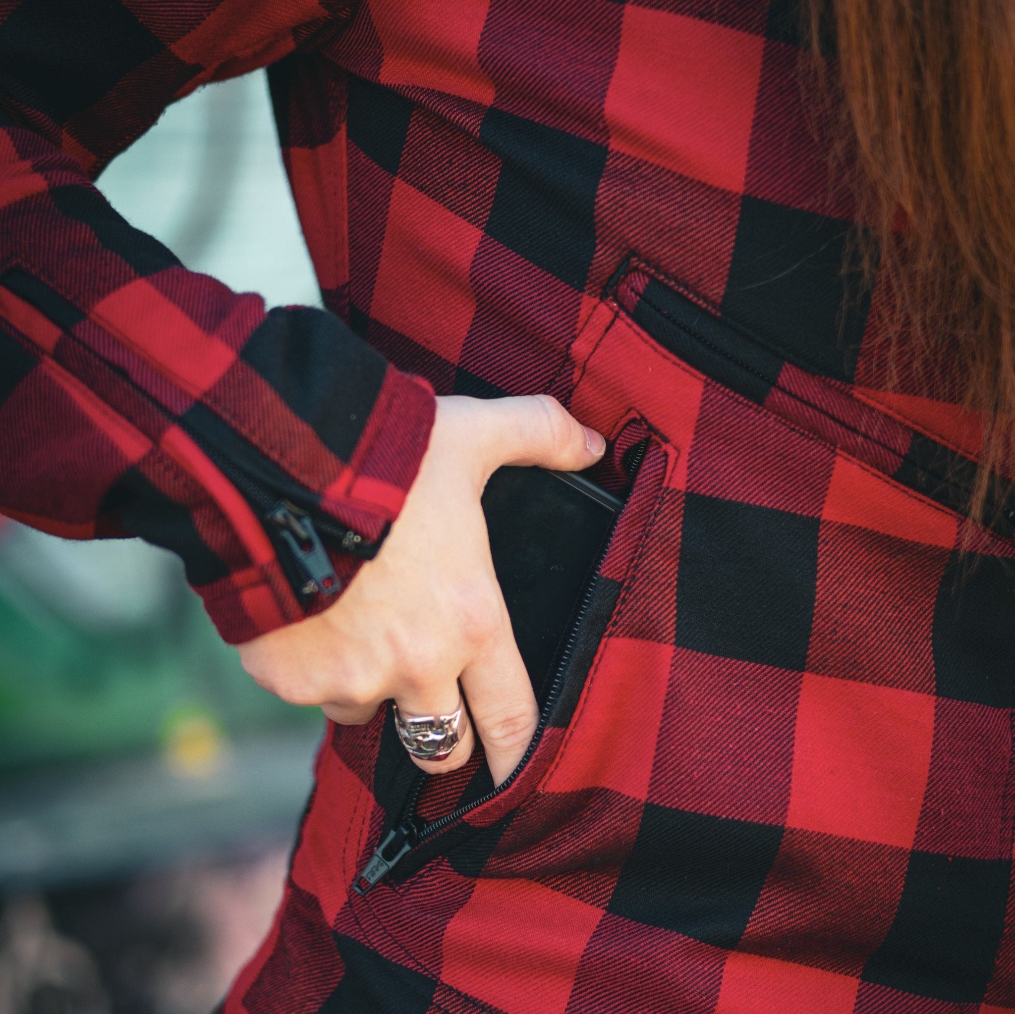 Close-up of a woman wearing a red and black flannel-style women&#39;s jacket, putting a phone in the side pocket.