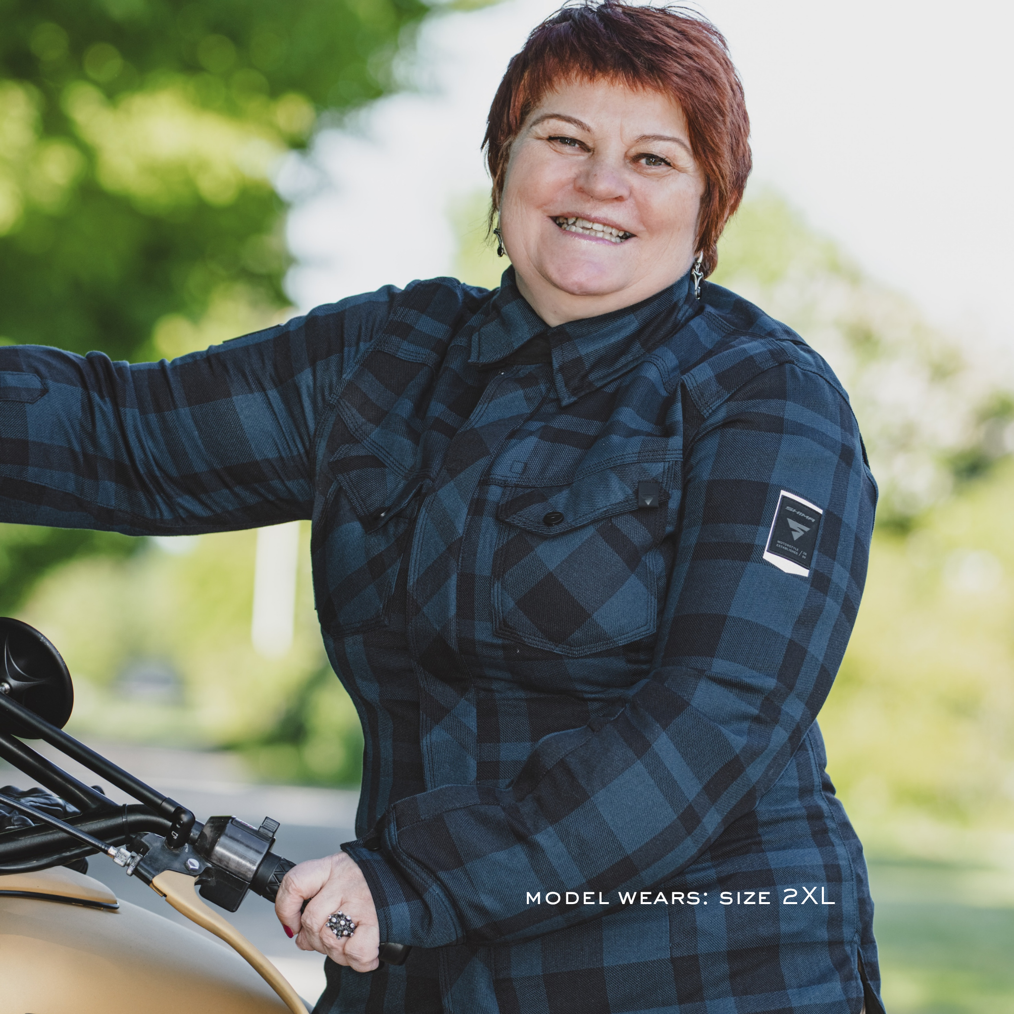 Person wearing a blue plaid shirt standing next to a motorcycle with a blurred green background