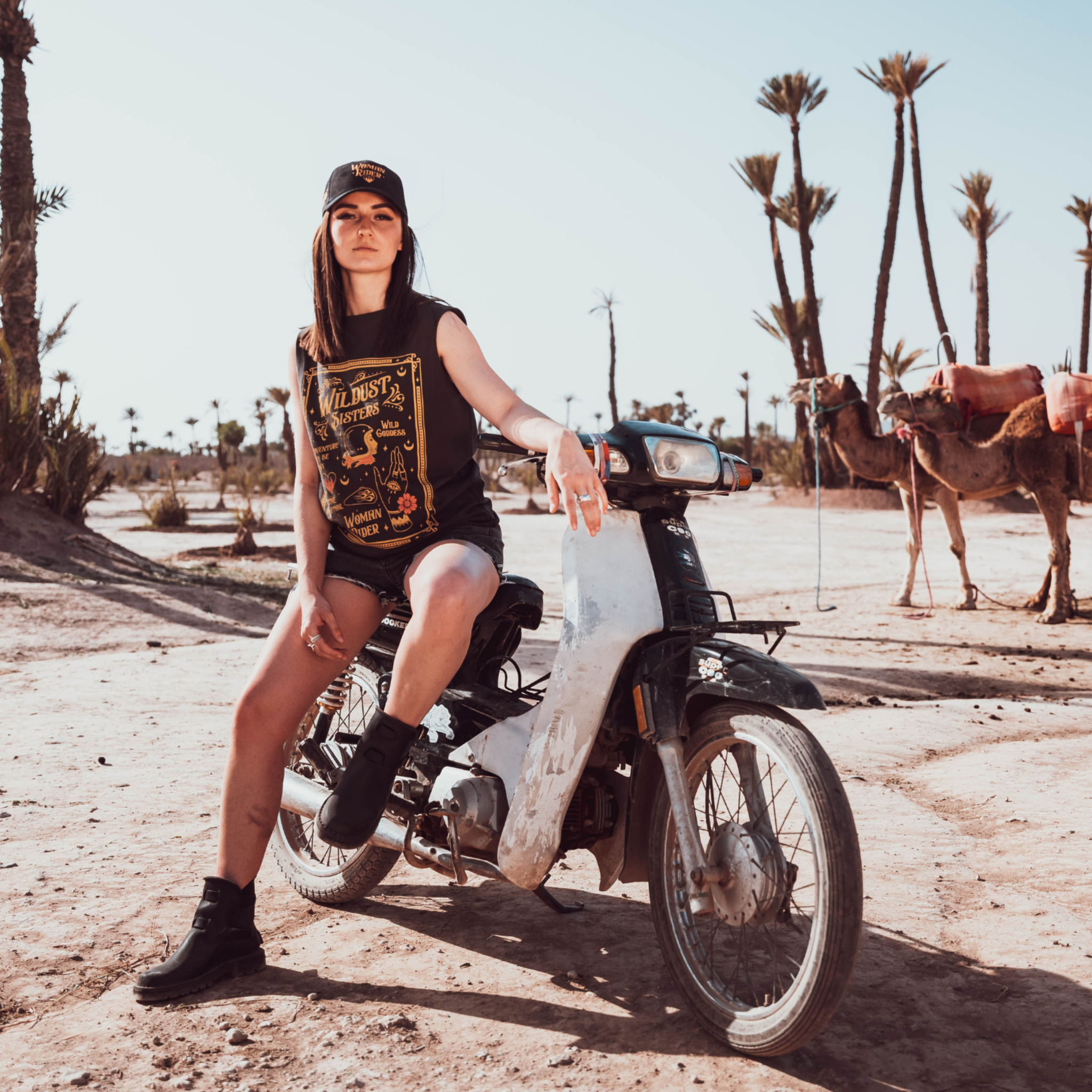 Woman sitting on a Motorcycle wearing a Black t-shirt with a woman wearing a Motorcycle helmet with the text "Wildust sister" 