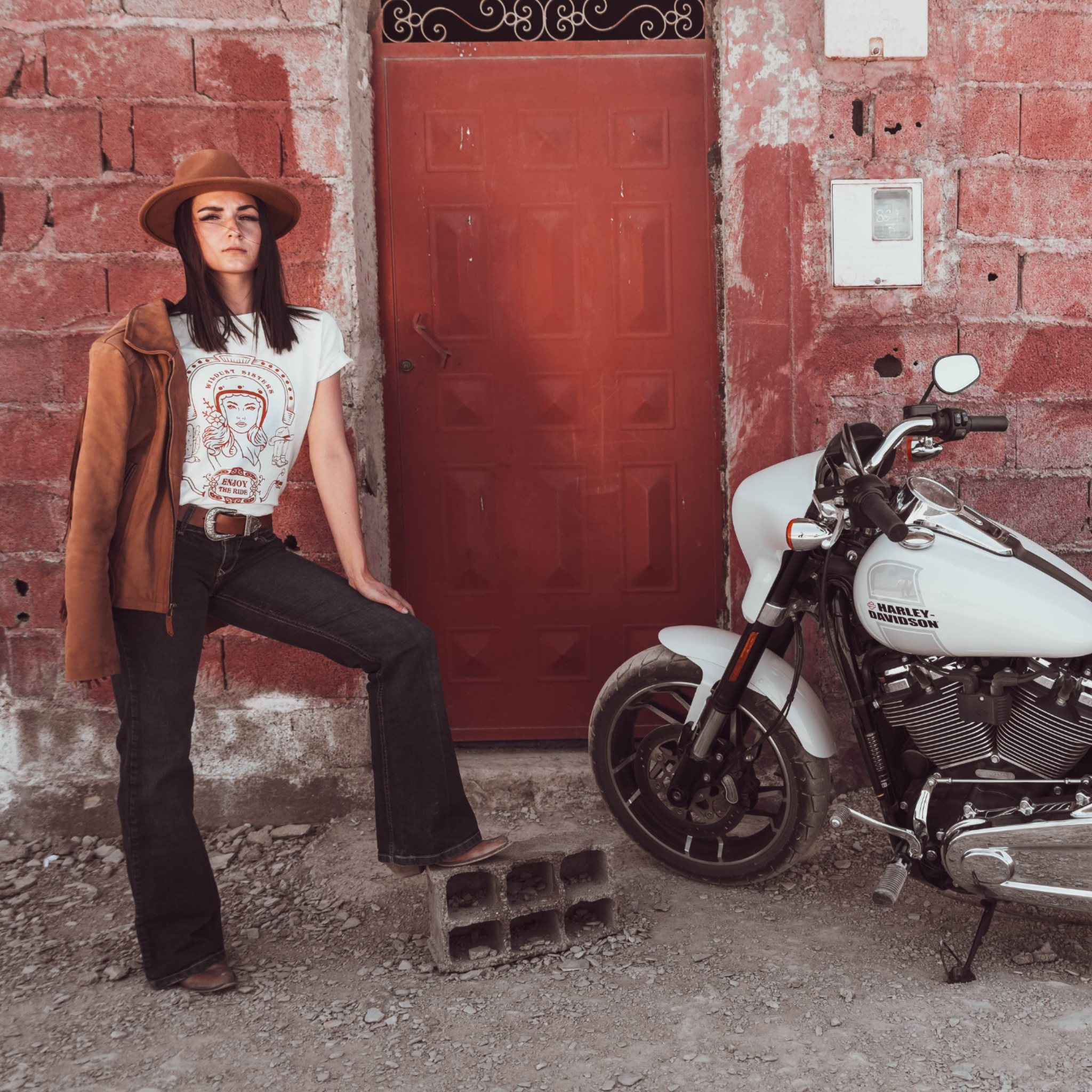 Woman standing next to a Motorcycle wearing a white T-shirt with a Woman wearing a Motorcyclehelmet print and the text &quot;Wild dust sister&quot; and &quot;Enjoy The ride&quot; in orange