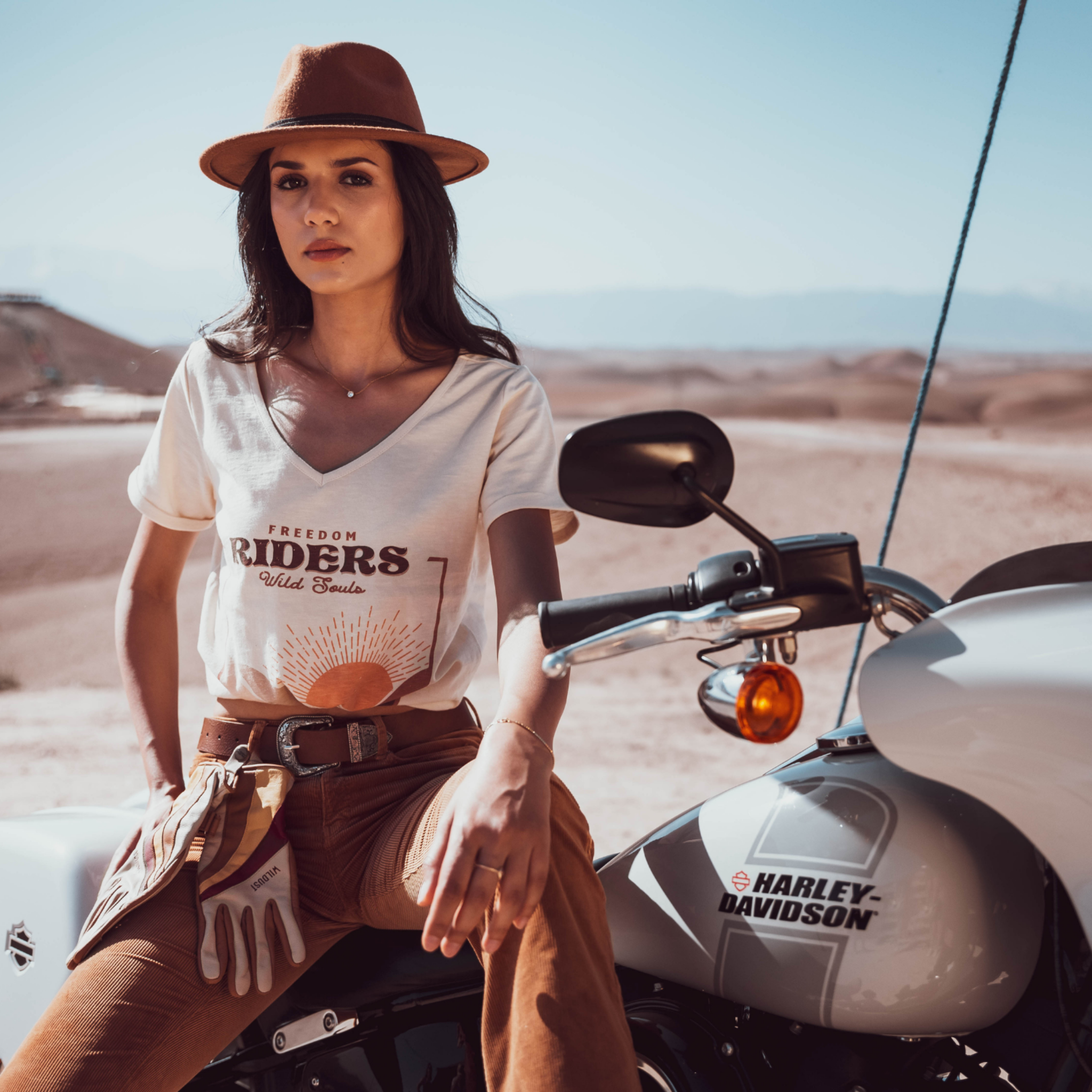 Woman sitting on a Motorcycle wearing a T-shirt with a sunset/sunrise print and the text "Freedom Riders Wild Souls" and "Wildust sisters"