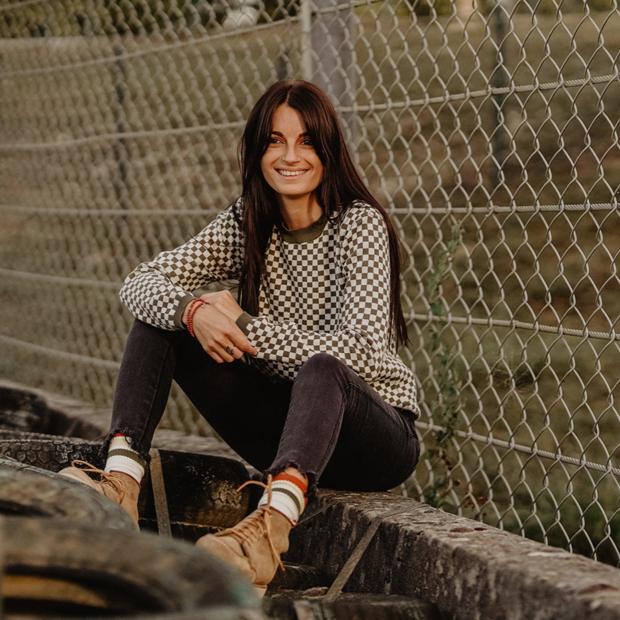 a young woman siting by the fence wearing a knitted khaki green and white jumper with chessboard motives  from Wildust