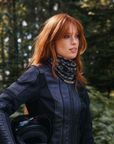 A woman with red hair wearing a black leather motorcycle jacket and a patterned neck gaiter, holding a black helmet in a forest setting.