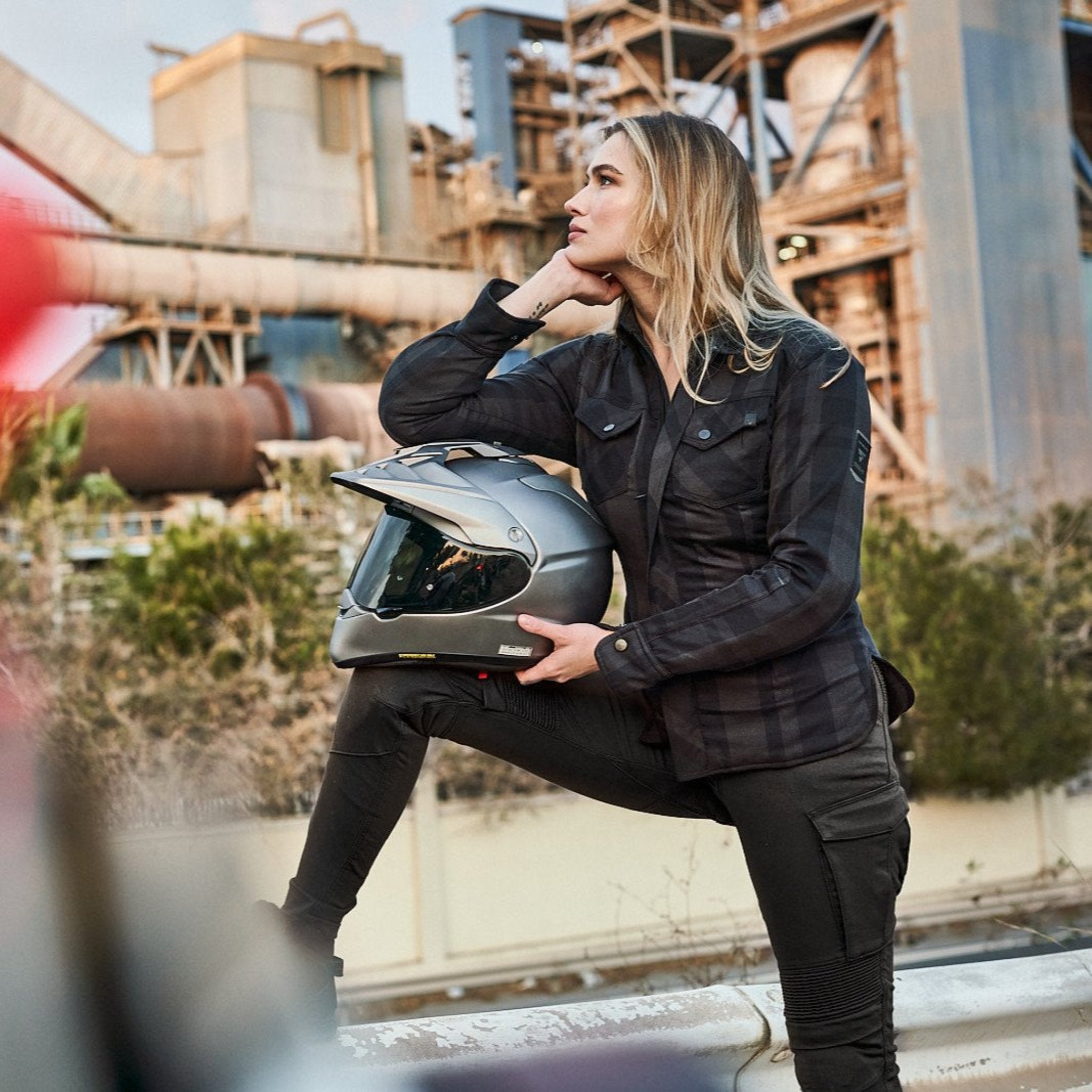 A blond young woman dreaming while holding her motorcycle helmet wearing Black and grey lumberjack style women's motorcycle shirt from Shima