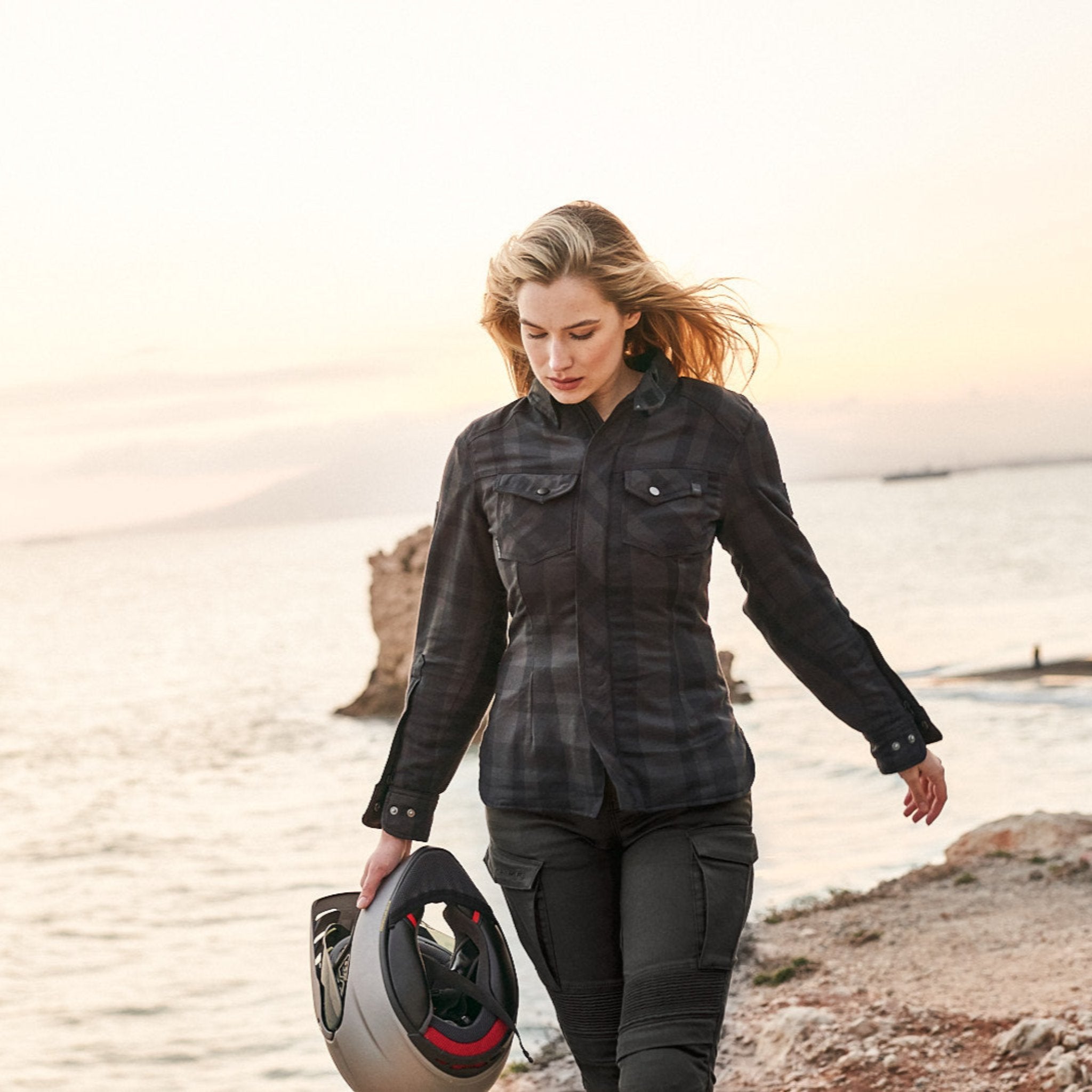 A blond young woman walking on a beach and carrying a motorcycle helmet wearing Black and grey lumberjack style women&#39;s motorcycle shirt from Shima