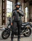 Person wearing a black leather jacket and helmet standing next to a motorcycle in an industrial setting.