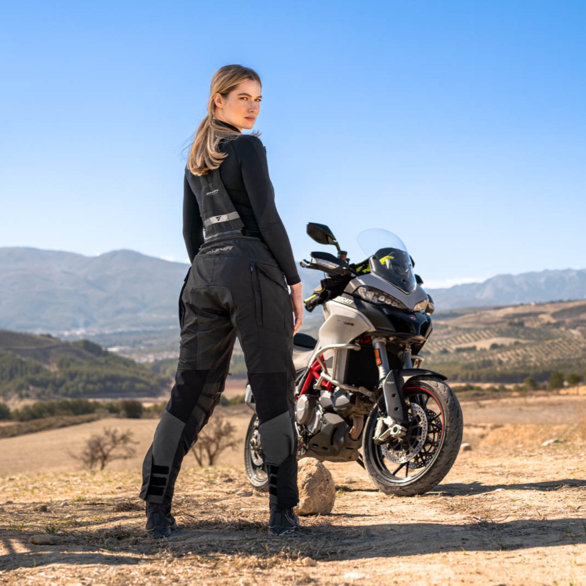 A woman standing by her motorcycle wearing Black SHIMA touring motorcycle pants for women