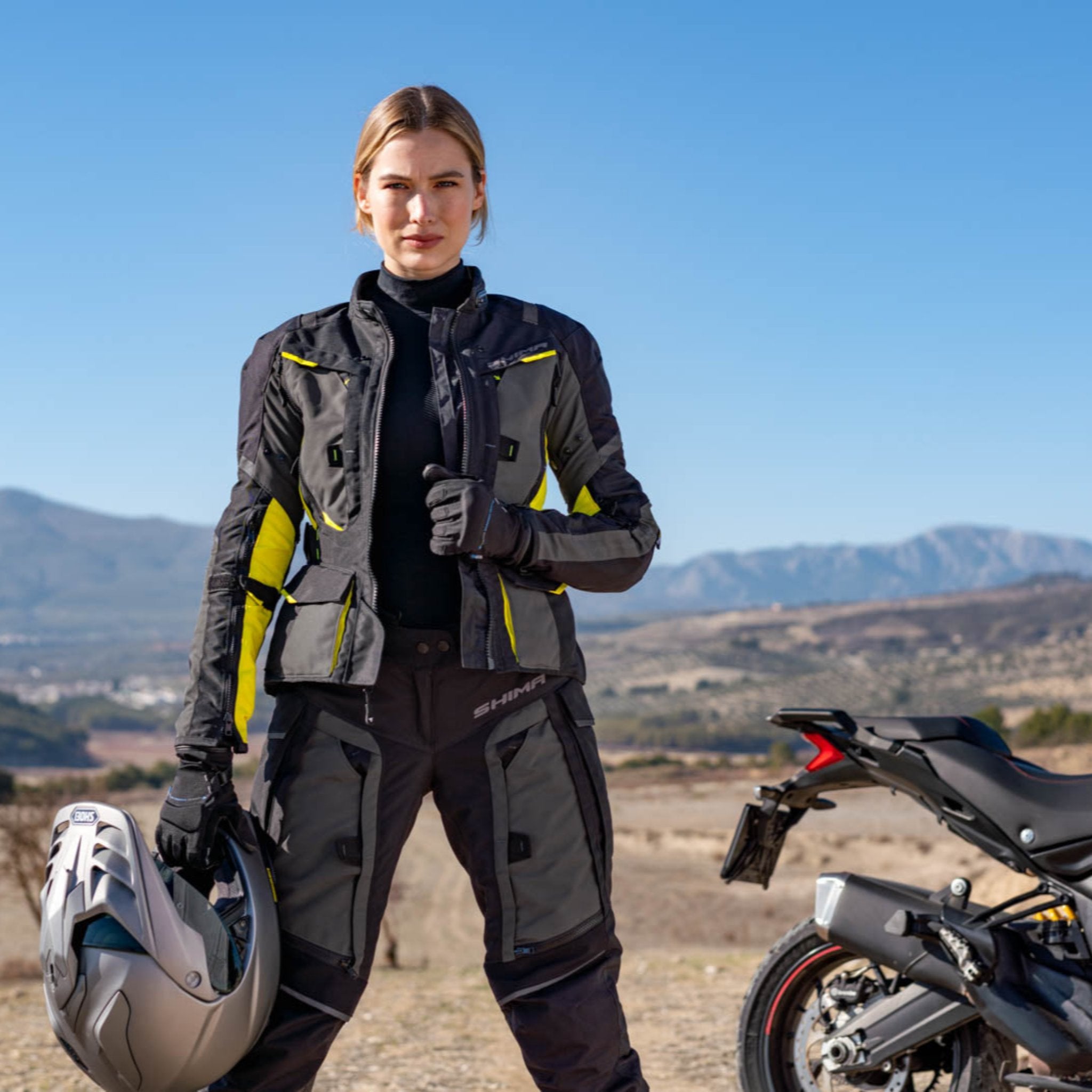 A young woman standing by her motorcycle wearing shima motorcycle touring jacket for women in black/ fluo