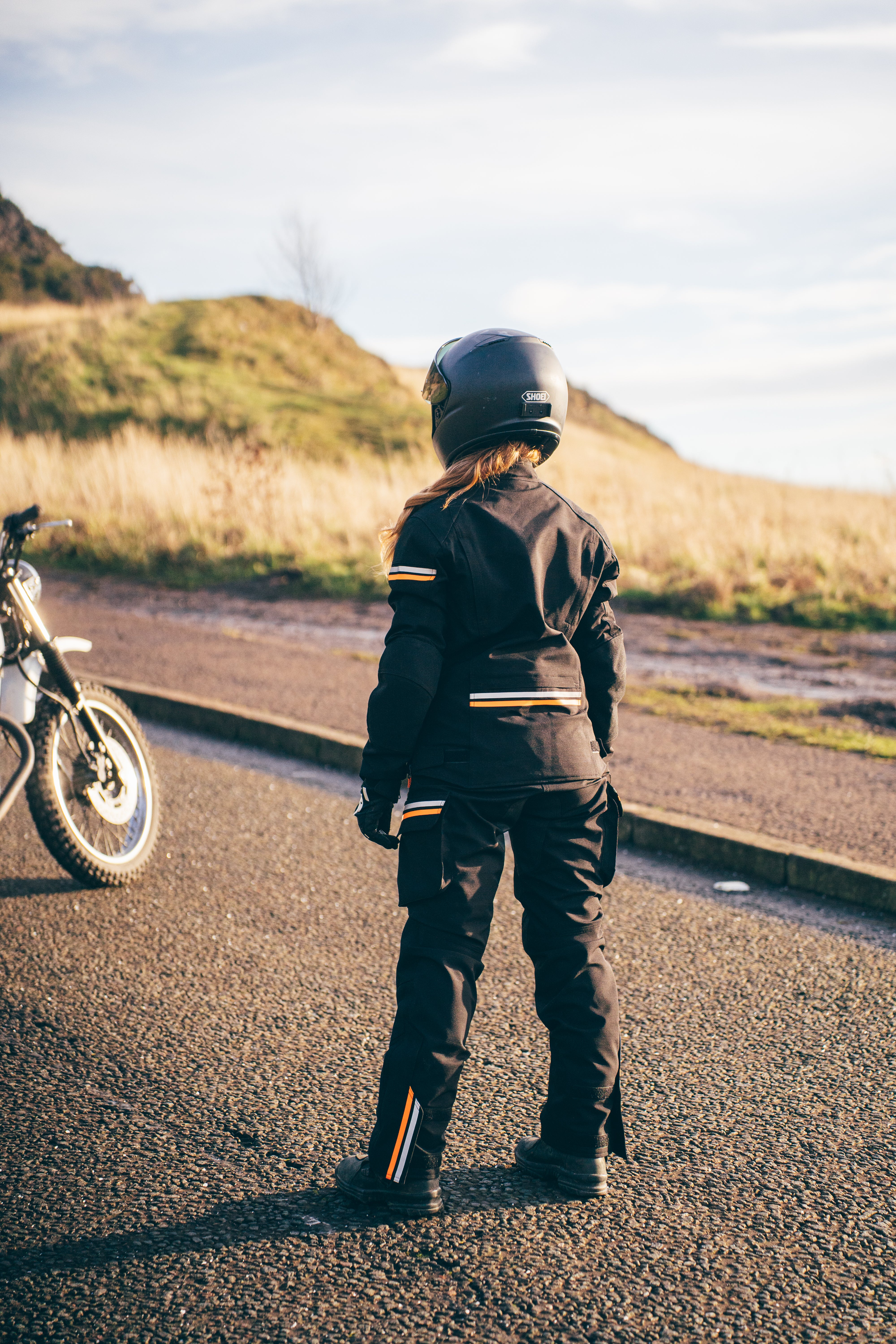 Person in motorcycle gear standing next to a motorcycle on a road with a scenic background
