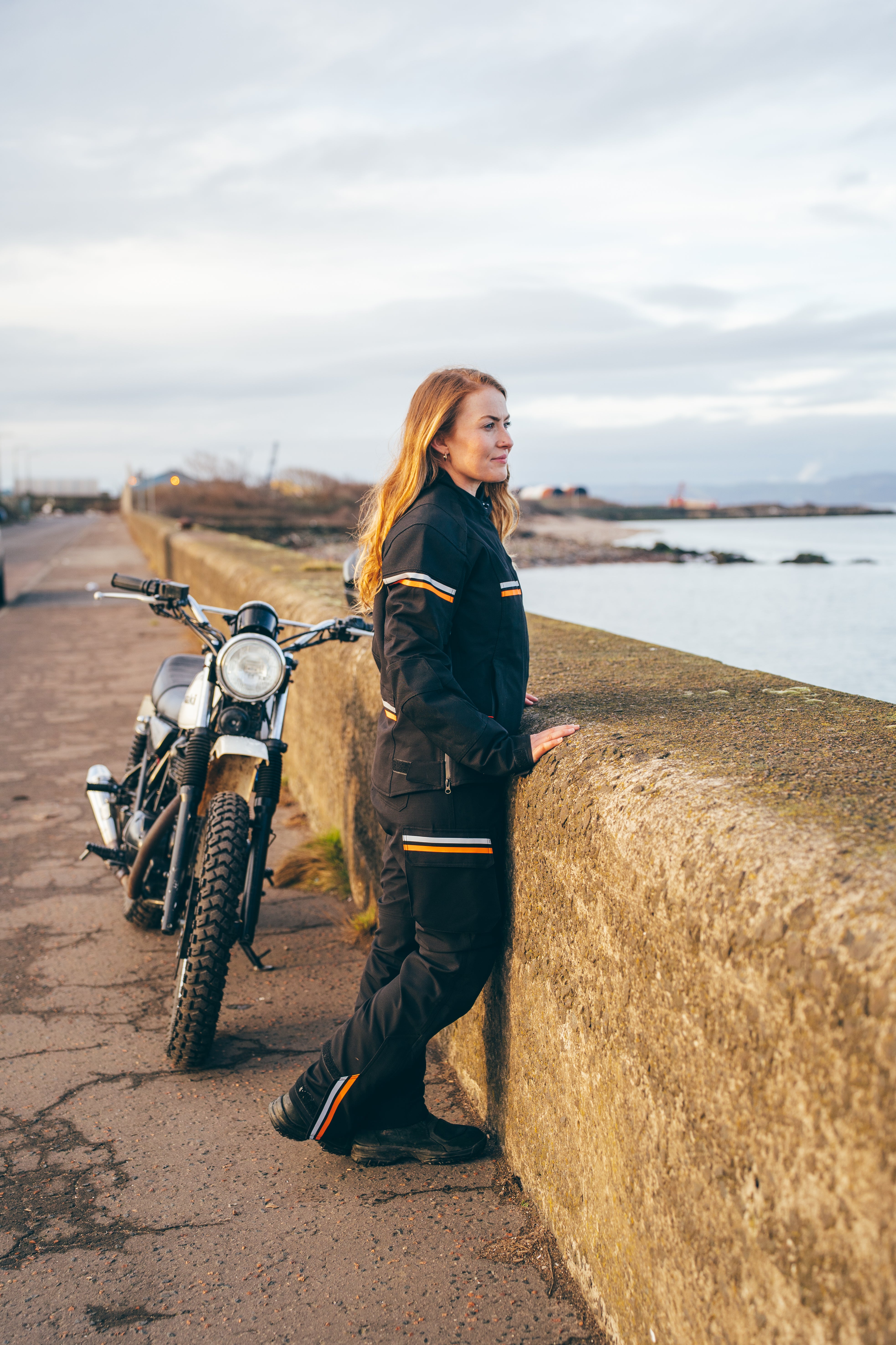 Person in motorcycle gear standing next to a motorcycle by a coastal road.