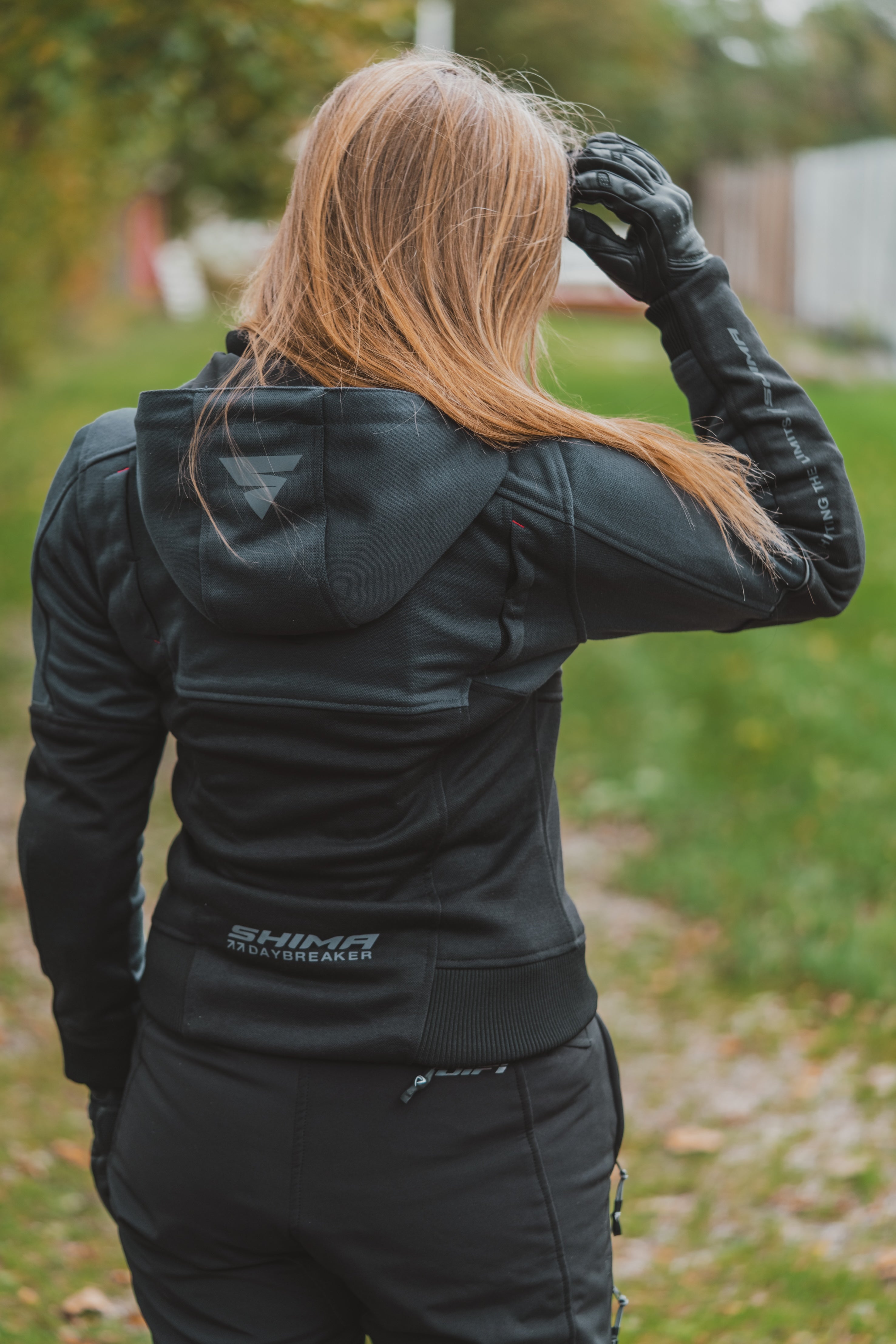 A woman viewed from behind wearing a black SHIMA insulated jacket and matching black pants, standing outdoors on a grassy path.