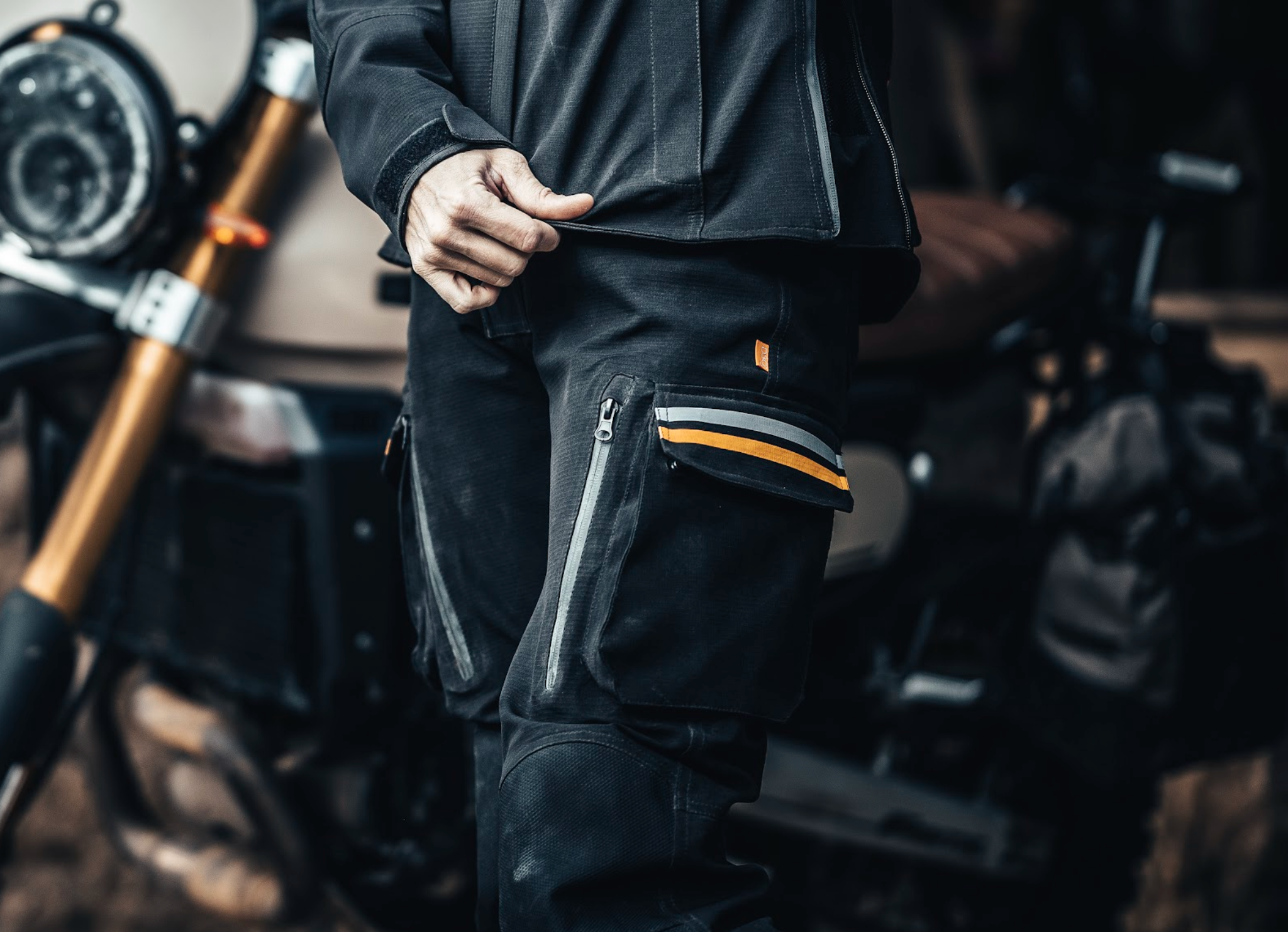 Person wearing black motorcycle pants with zippers and reflective stripes, standing next to a motorcycle.