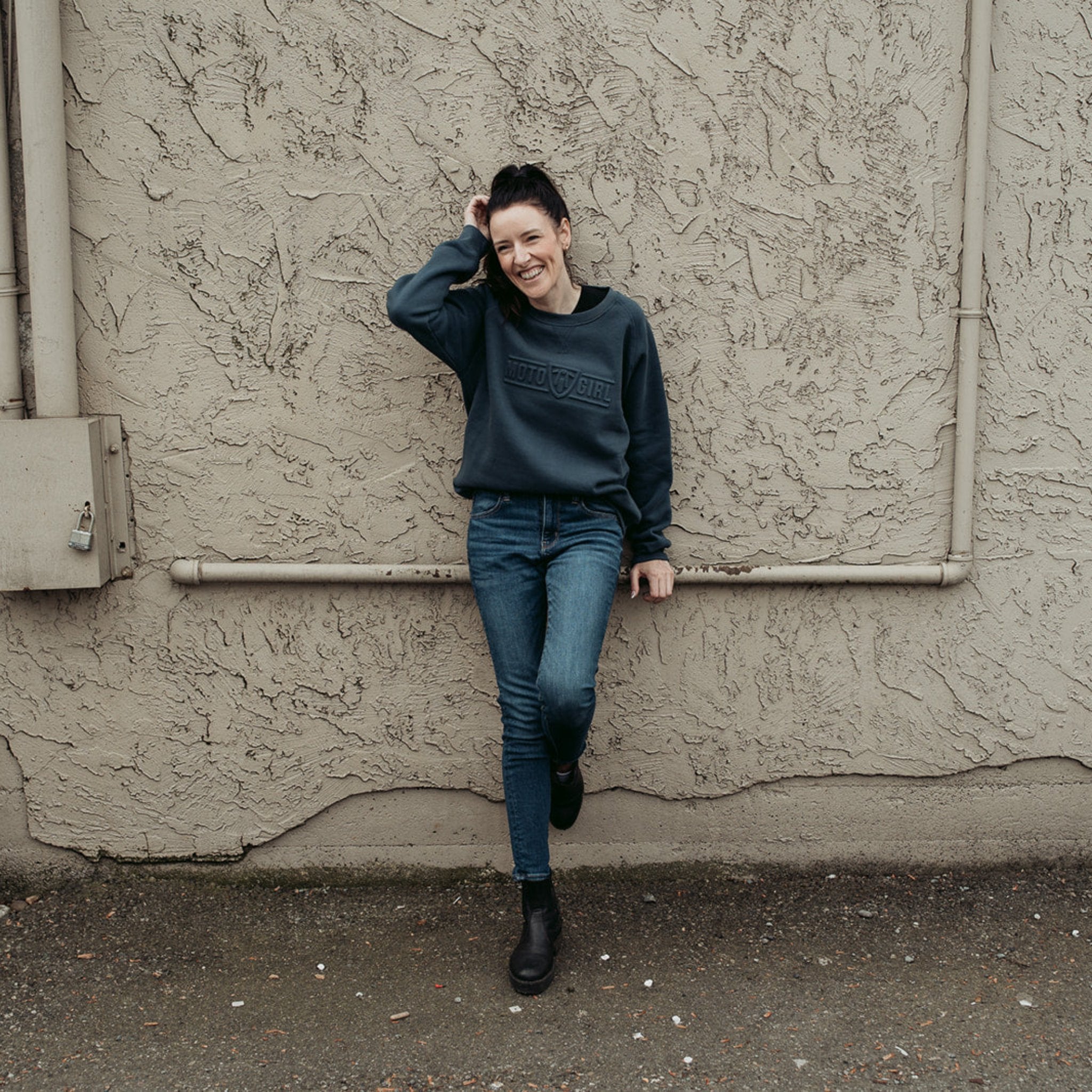 A young woman standing by a wall and wearing  dark blue colour lady sweatshirt with Moto Girl 3D logo