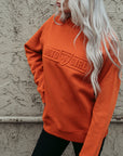 A young woman standing by a wall and wearing orange colour lady sweatshirt with Moto Girl 3D logo