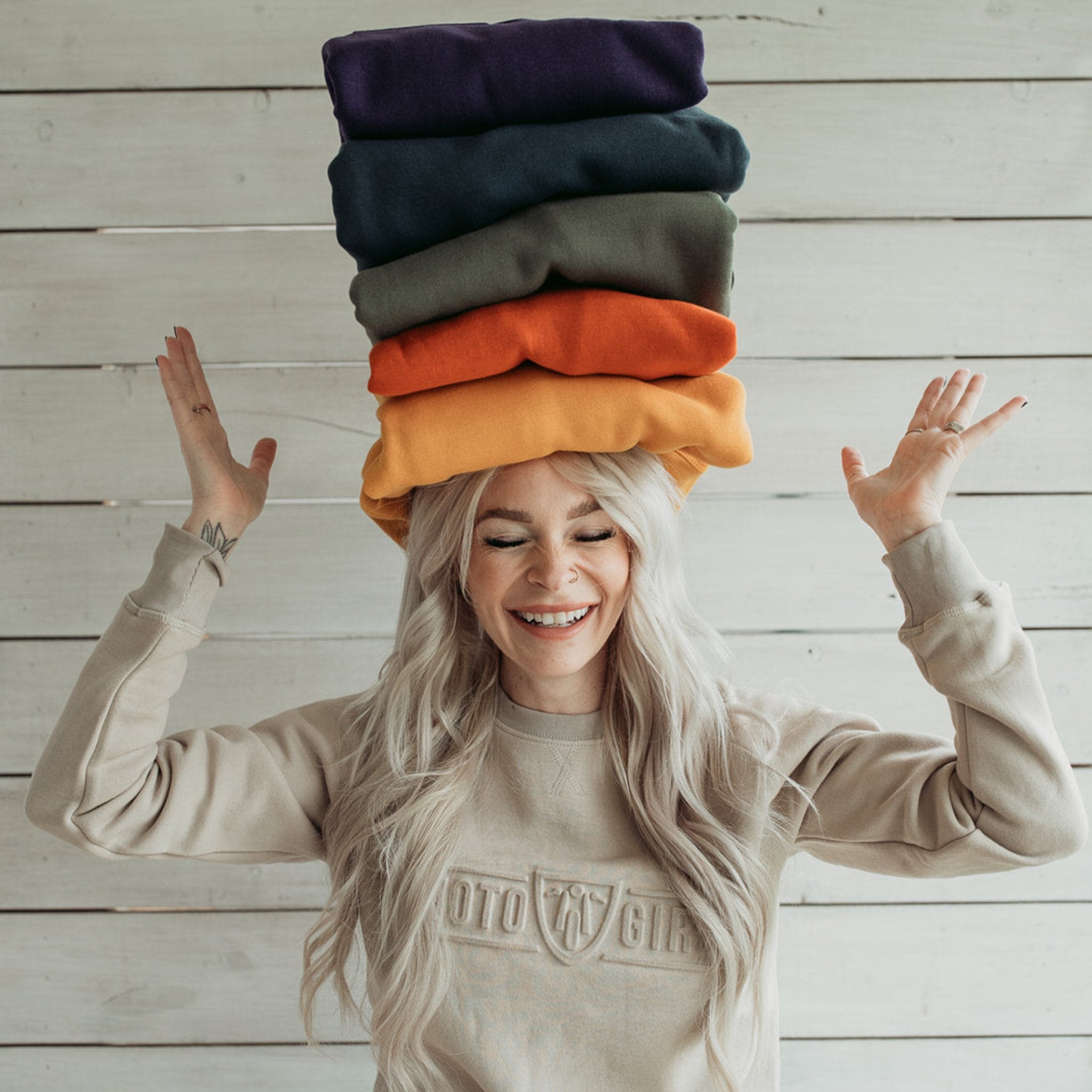 a young woman wearing Sand colour lady sweatshirt with Moto Girl 3D logo and holding 5 colourful sweatshirts on her head