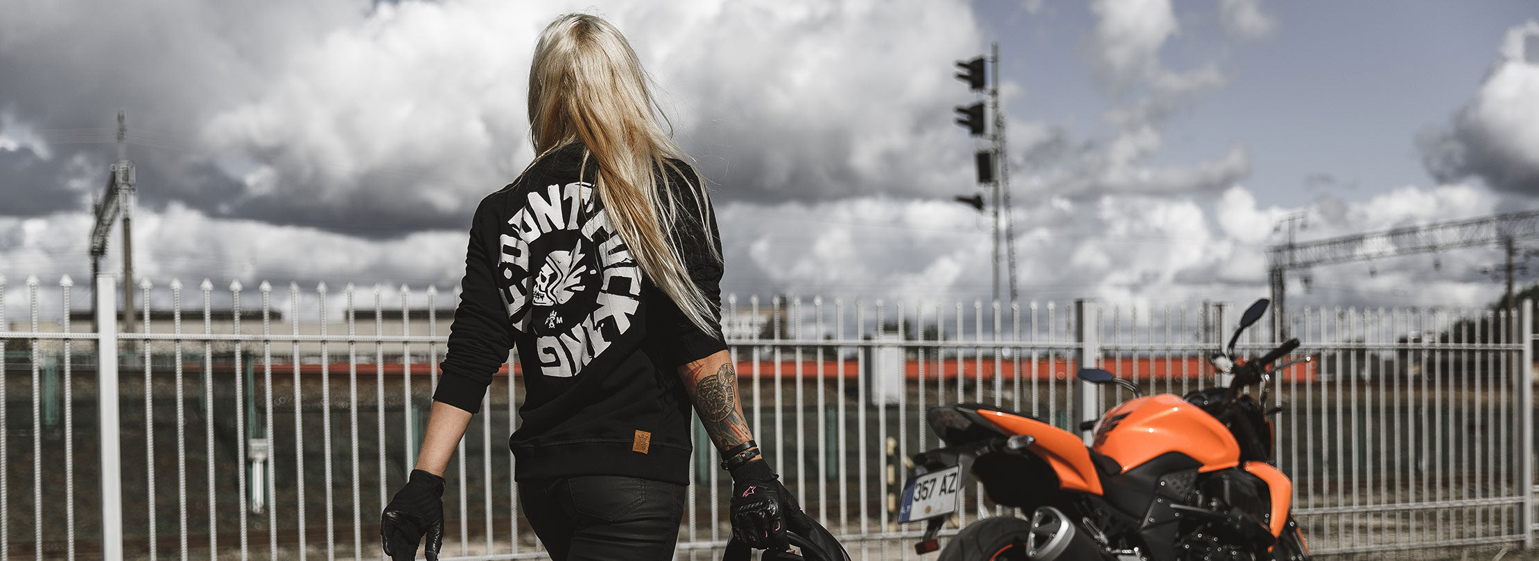A blond woman walking towards the orange motorcycle wearing Pando Moto black sweatshirt with while &quot;Don&#39;t Fucking Die&quot; motive on the back