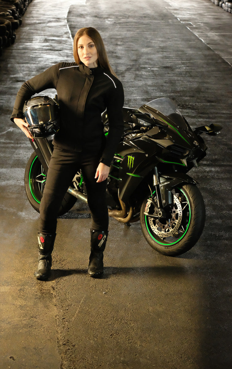 A young woman standing by the motorcycle wearing black motorcycle protective clothes