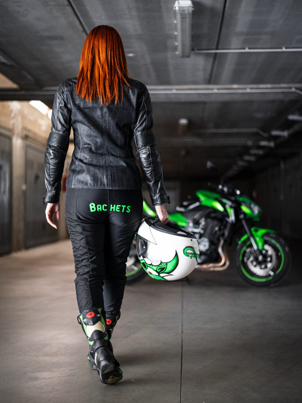 A red hair woman walking towards her motorcycle wearing black and red women's motorcycle leggings from exagon66