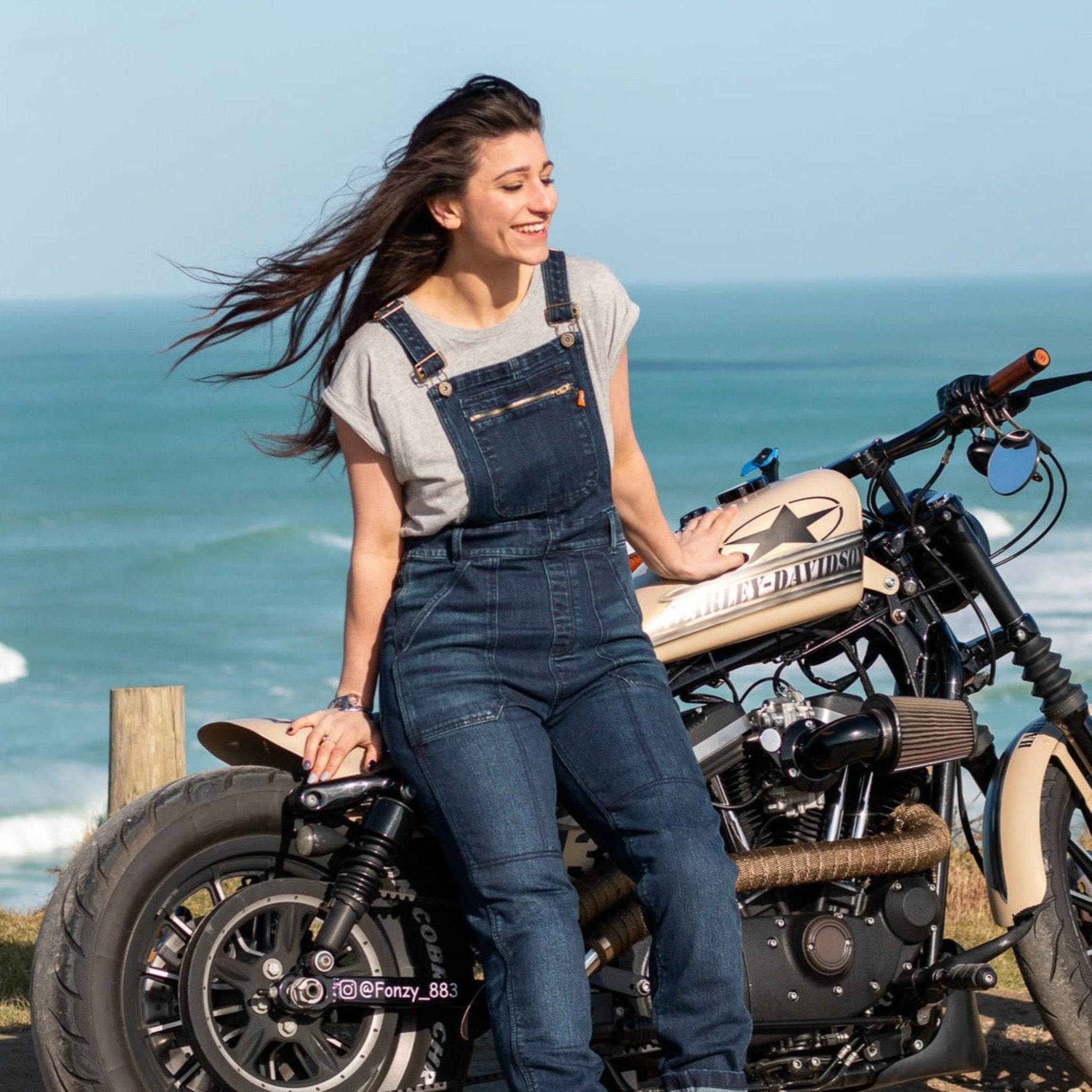 A woman by the beach with her motorcycle wearing blue women&#39;s motorcycle overall from Moto Girl with zipper pocket detail on the chest