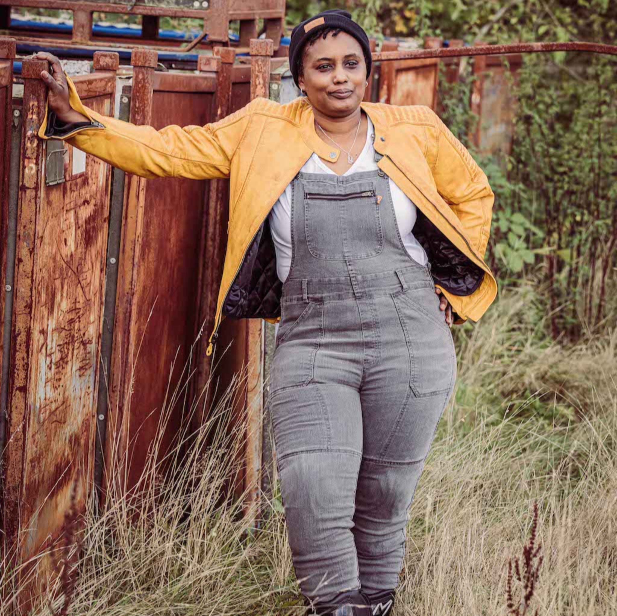 A woman wearing grey motorcycle overall and yellow leather jacket standing by the metal container