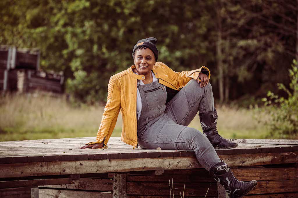 A woman wearing grey motorcycle overall and yellow leather jacket sitting on the wooden bridge 
