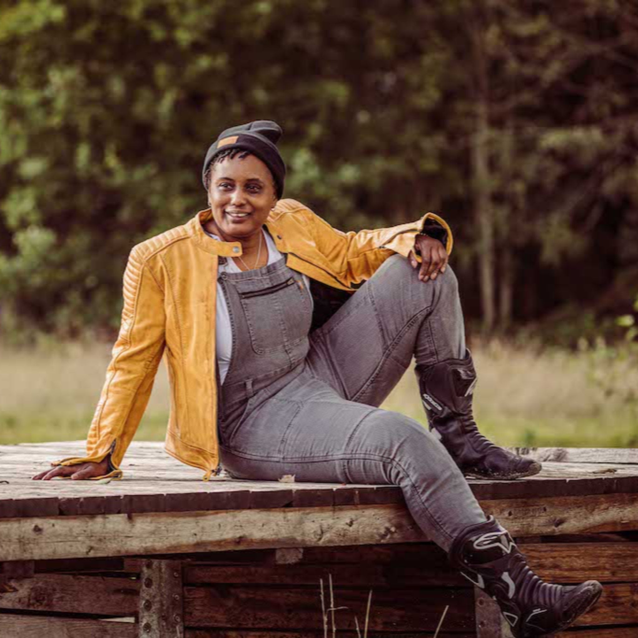 A woman wearing grey motorcycle overall and yellow leather jacket sitting on the wooden bridge 