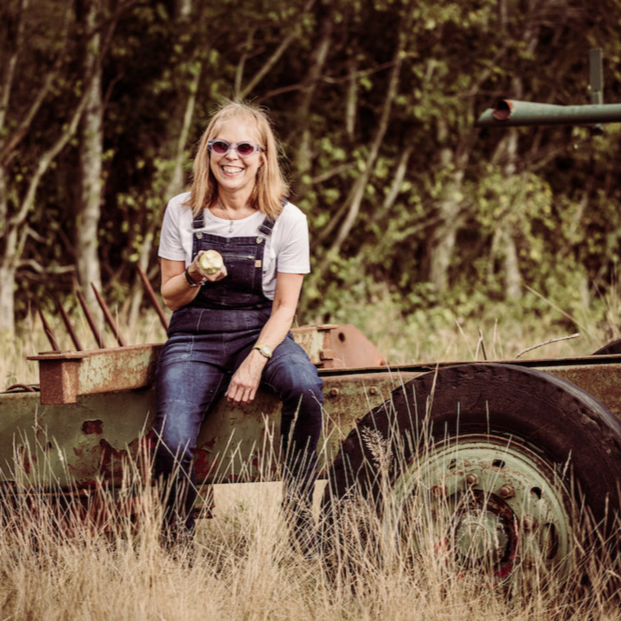 Blonde woman with blue overalls and a white t-shirt sitting on the back of a tractor