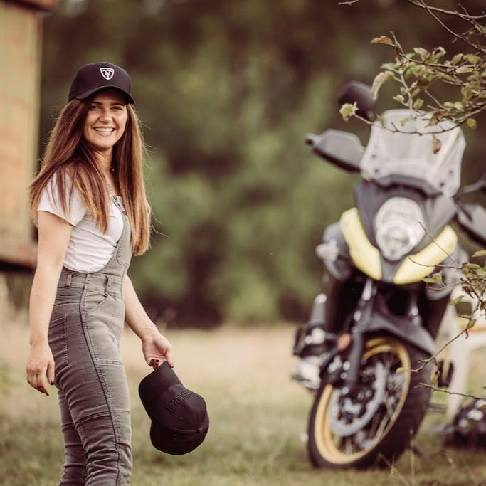 A woman wearing white tshirt, moto girl cap and grey motorcycle overall standing close to a motorcycle