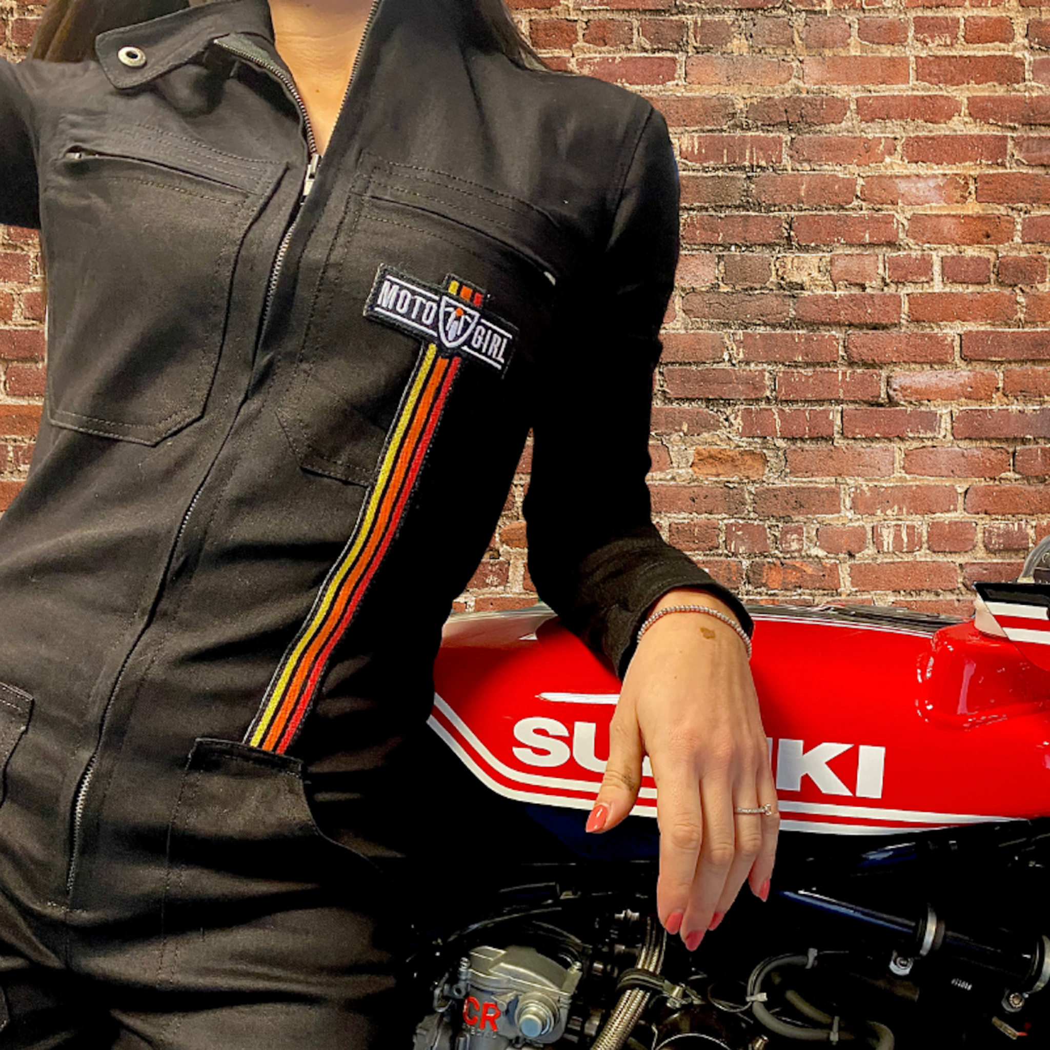 A women leaning on a red Suzuki motorcycle wearing Black garage suit for women from MotoGirl with red, orange and yellow stripe details