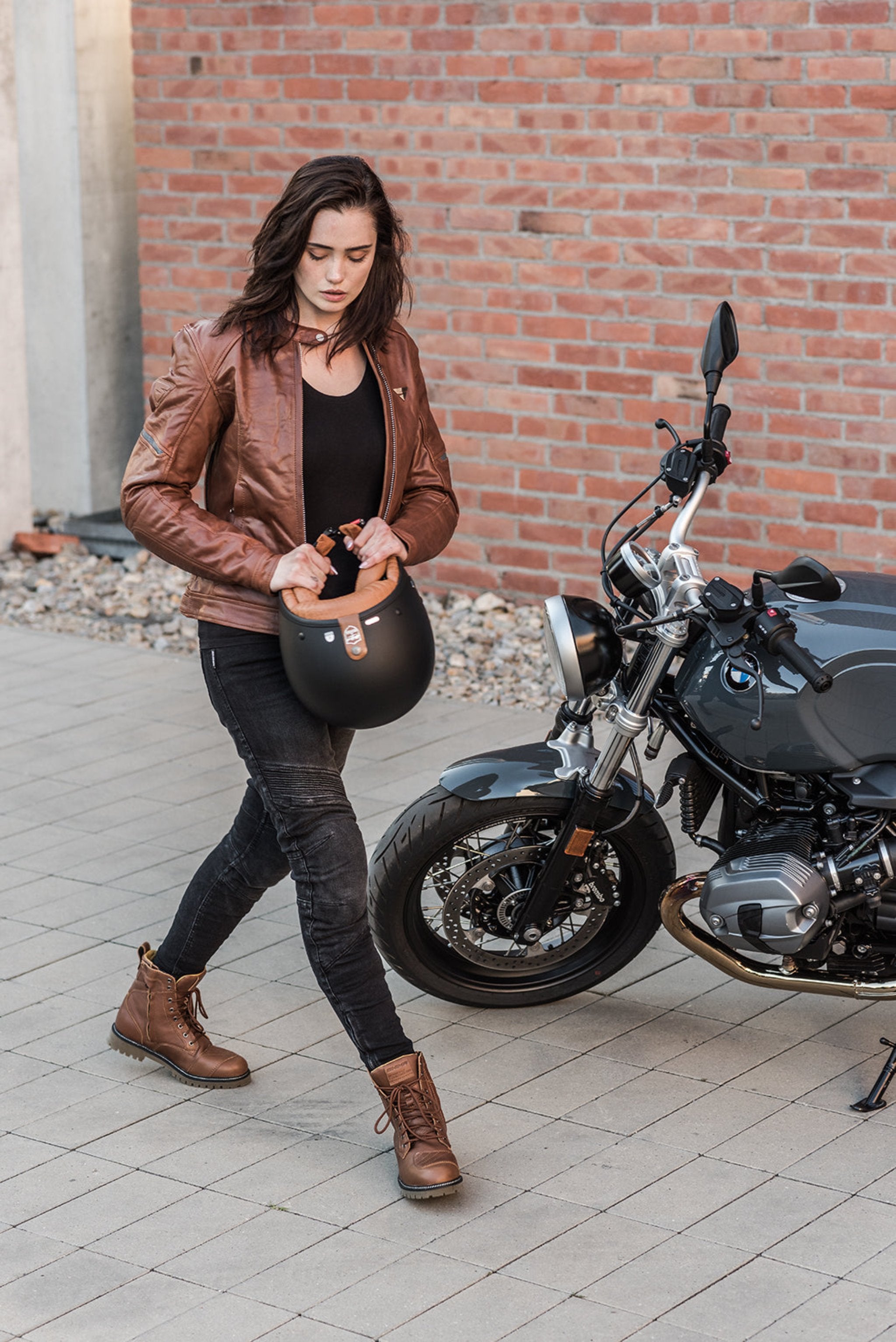 A young woman by a motorcycle wearing brown motorcycle leather jacket and brown women motorcycle boots from Shima